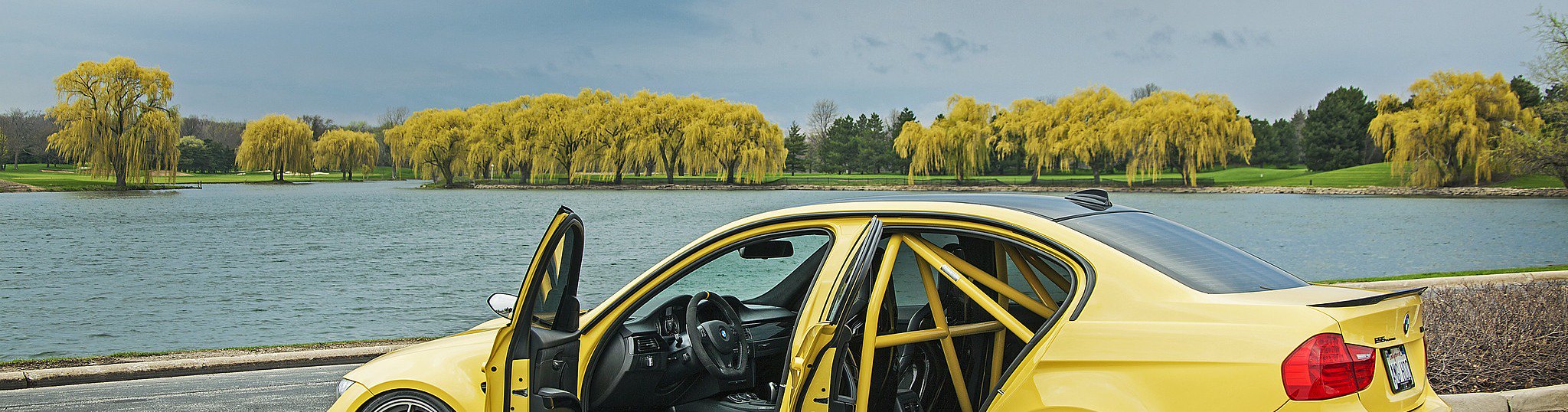 Dakar Yellow BMW E90 M3 sedan with doors open showing yellow-matched roll cage parked lakeside on BBS forged wheels