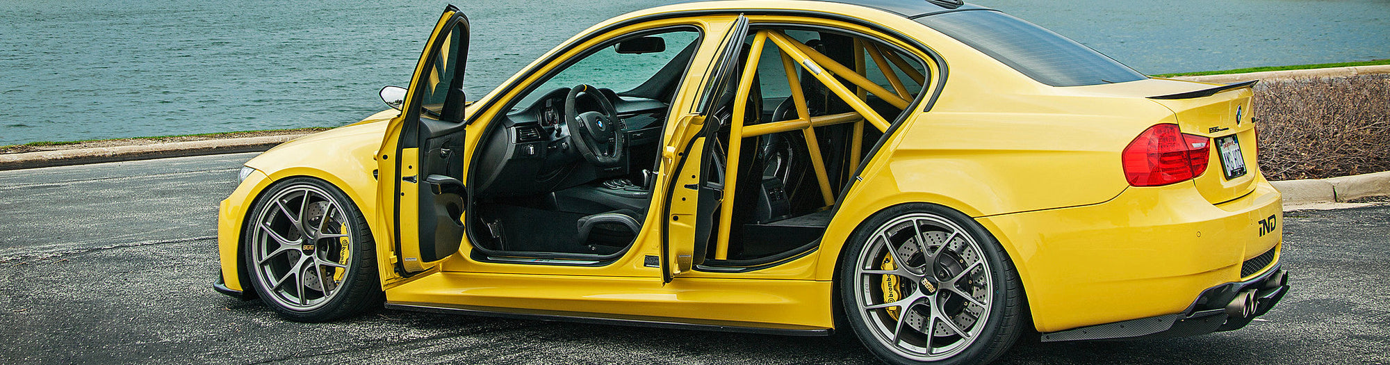 Banner hero image showing IND’s Dakar Yellow BMW E90 M3 from the side with the front and rear doors open revealing the matching Dakar Yellow roll bar and custom interior details.