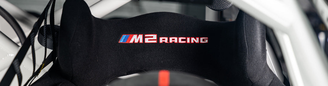 Close-up of the BMW M2 Racing Sabelt racing seat headrest featuring the embroidered M2 Racing logo within the motorsport cockpit.