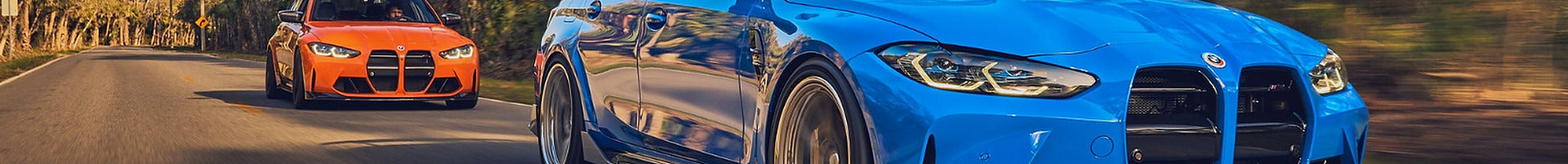 Blue BMW G80 M3 Competition leading an orange G80 M3 down a tree-lined road