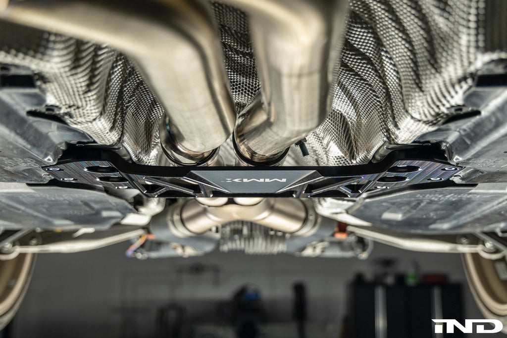 MMX Performance billet aluminum underbody chassis brace installed on a BMW G8X M3 and M4, providing an additional 15mm of exhaust clearance while increasing chassis rigidity for aftermarket exhaust systems.