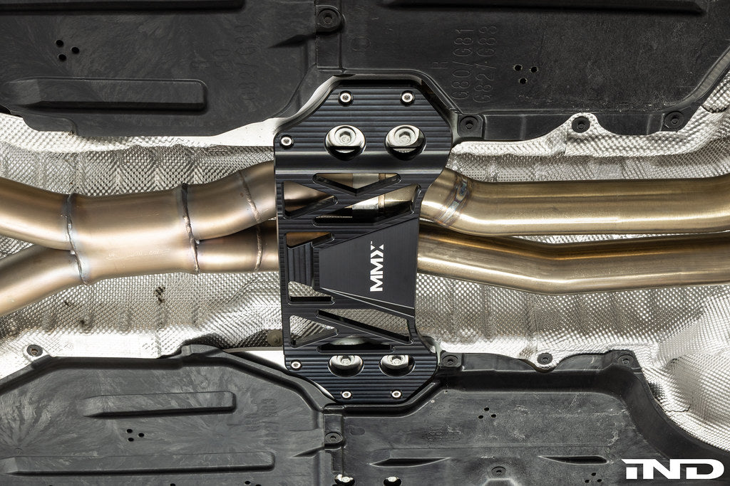 MMX Performance billet aluminum underbody chassis brace installed on a BMW G8X M3 and M4, providing an additional 15mm of exhaust clearance while increasing chassis rigidity for aftermarket exhaust systems.