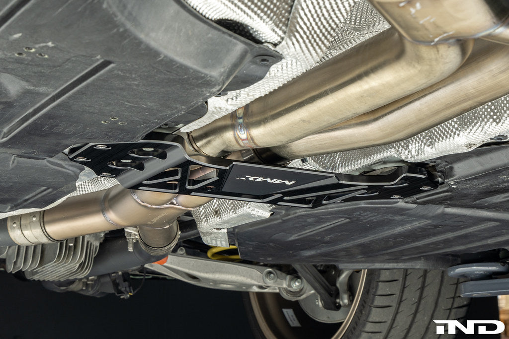 MMX Performance billet aluminum underbody chassis brace installed on a BMW G8X M3 and M4, providing an additional 15mm of exhaust clearance while increasing chassis rigidity for aftermarket exhaust systems.