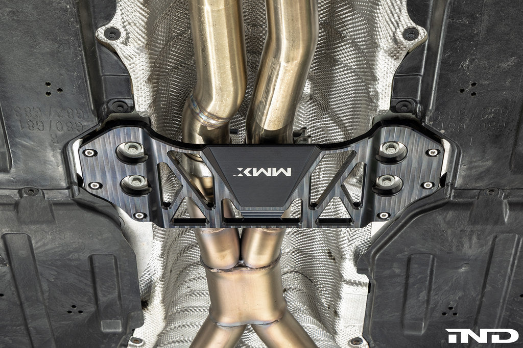 MMX Performance billet aluminum underbody chassis brace installed on a BMW G8X M3 and M4, providing an additional 15mm of exhaust clearance while increasing chassis rigidity for aftermarket exhaust systems.