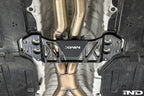 MMX Performance billet aluminum underbody chassis brace installed on a BMW G8X M3 and M4, providing an additional 15mm of exhaust clearance while increasing chassis rigidity for aftermarket exhaust systems.