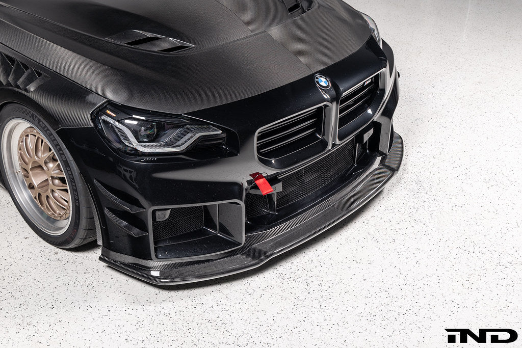 Alpha-N Carbon Corse front splitter installed on a Black Sapphire BMW G87 M2, enhancing the front-end appearance with pre-preg carbon fiber construction and aggressive styling.