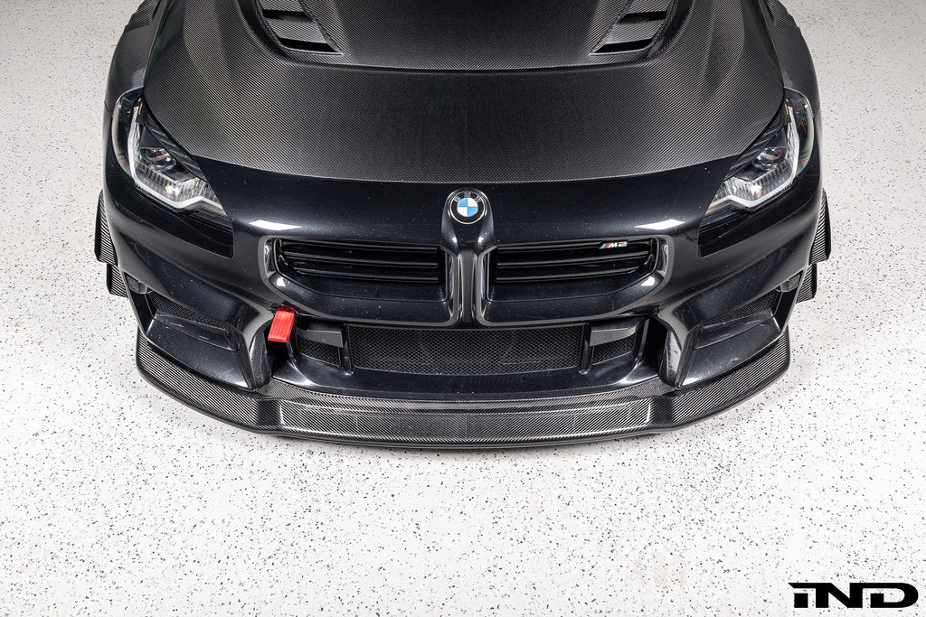 Alpha-N Carbon Corse front splitter installed on a Black Sapphire BMW G87 M2, enhancing the front-end appearance with pre-preg carbon fiber construction and aggressive styling.