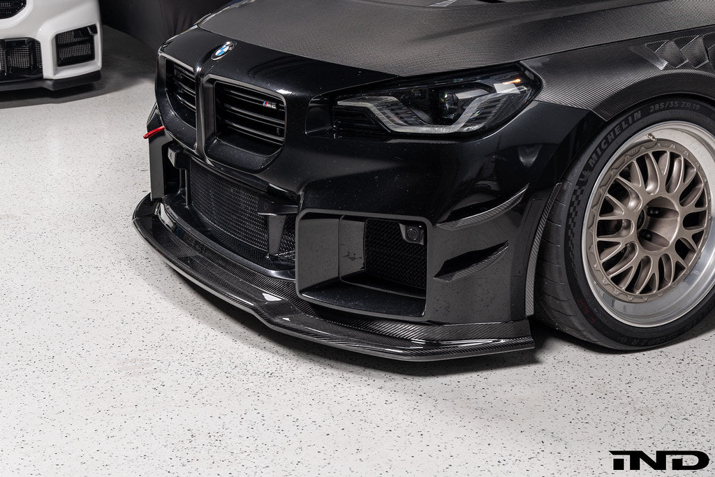 Alpha-N Carbon Corse front splitter installed on a Black Sapphire BMW G87 M2, enhancing the front-end appearance with pre-preg carbon fiber construction and aggressive styling.