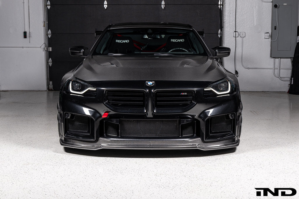 Alpha-N Carbon Corse front splitter installed on a Black Sapphire BMW G87 M2, enhancing the front-end appearance with pre-preg carbon fiber construction and aggressive styling.