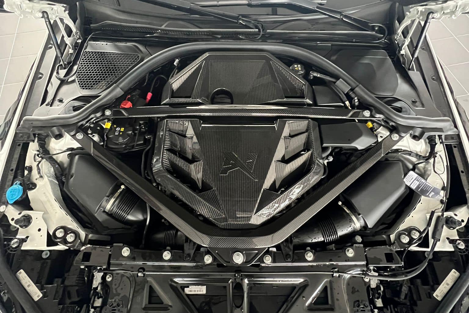 Alpha-N G8X S58 carbon engine cover installed on BMW M2/M3/M4, exterior view showing sleek black carbon finish.