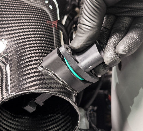Close-up of the Eventuri E36/7 Z3M Hybrid Carbon Airbox CNC-machined MAF boss during installation, showing a technician wearing a black glove securing the factory MAF sensor to the carbon fiber snorkel assembly.