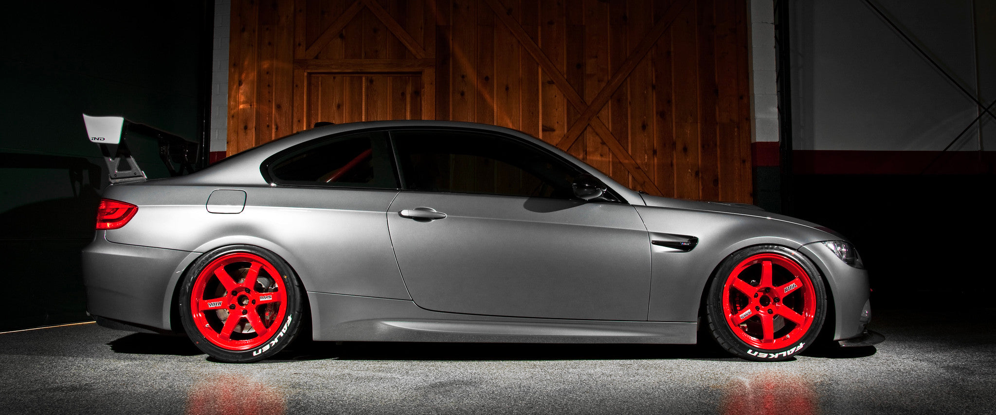 Side profile view of an IND BMW E92 M3 finished in Space Grey with red Volk TE37 wheels and a Varis Japan rear wing, photographed in a dark warehouse as a homepage hero image.