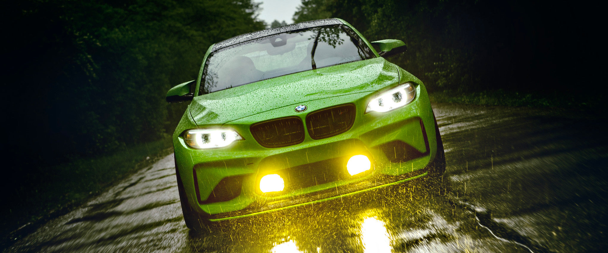 IND BMW F87 M2 in Gelbgrun featured as a homepage hero image, shown from the front in rainy conditions with yellow motorsport fog lights illuminated.