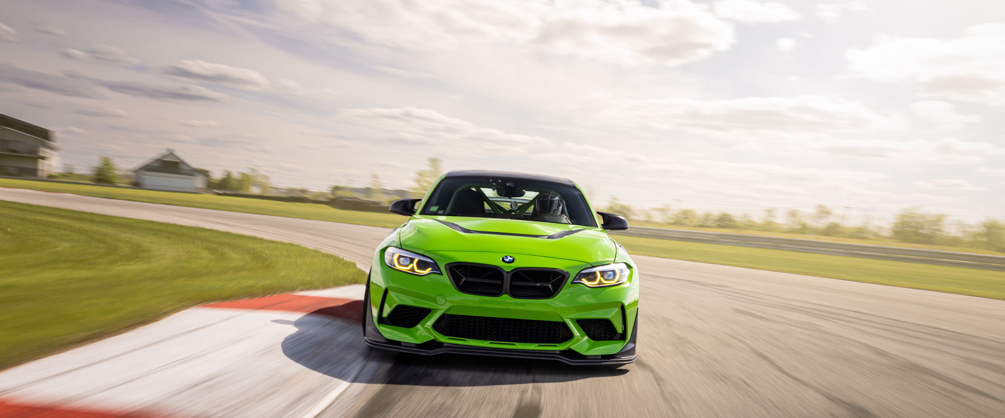 Front view of IND's BMW F87 M2 on track at the apex of a corner, passing over red and white curbing during a high-performance driving session, featured as a homepage hero image.