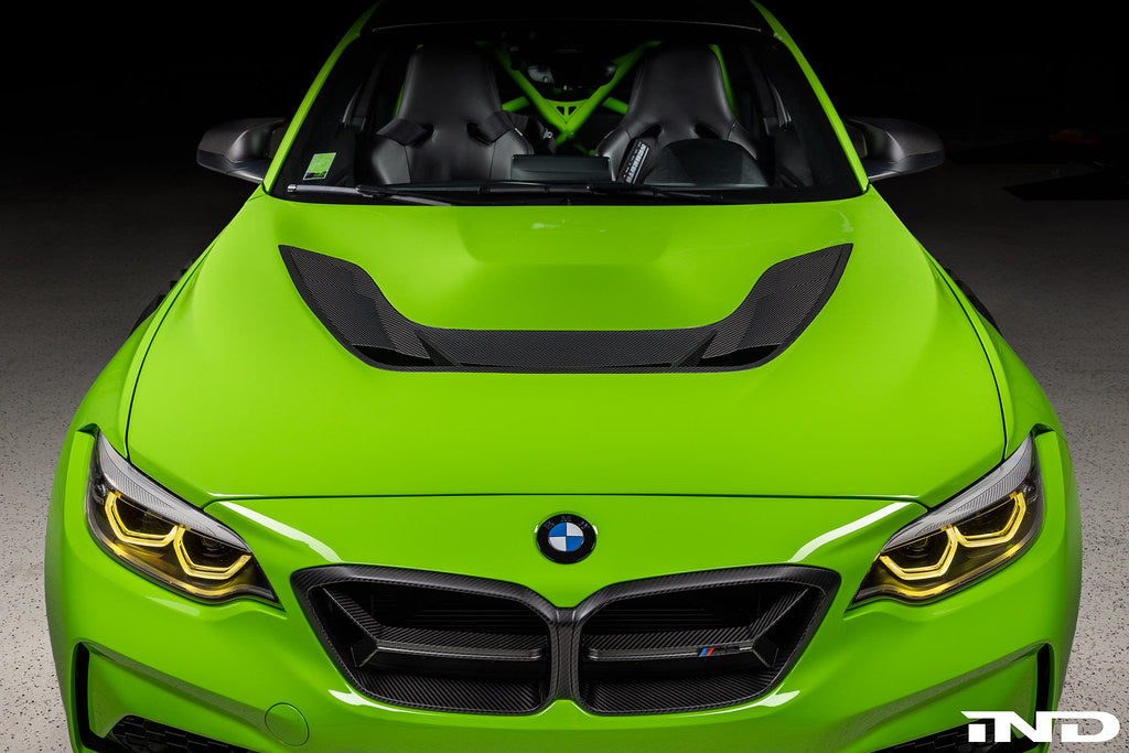 Alpha N F87 M2 Carbon Corse vented hood installed on the exterior of a car, showcasing its lightweight, aerodynamic design and vent details.