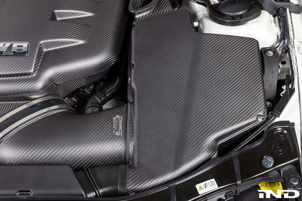 Close-up overhead installed view of Eventuri E9X M3 S65 matte black carbon airbox lid with optional intake and plenum in engine bay
