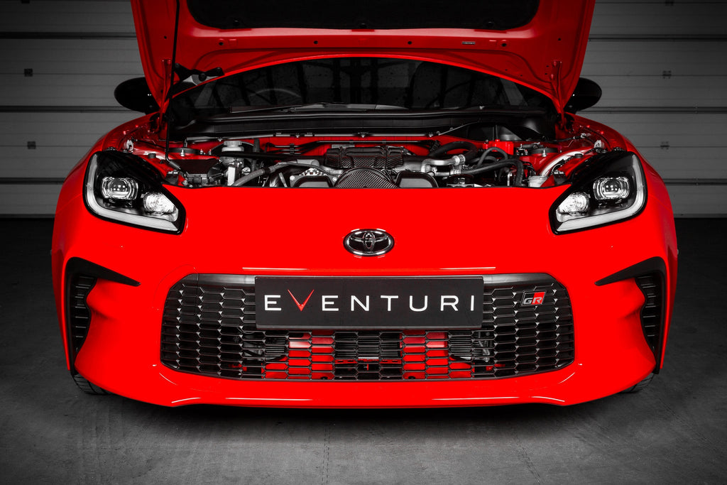 Eventuri Toyota GR86 black carbon intake system installed, showing the performance air intake with engine cover in a car engine bay.