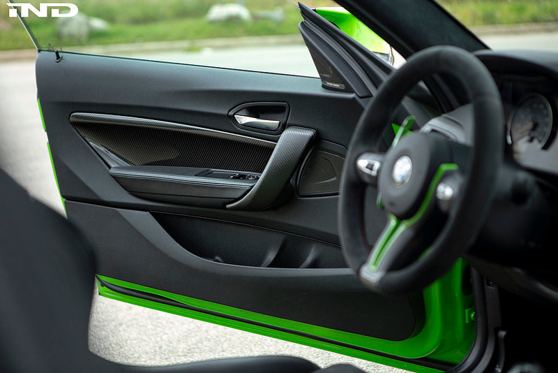 Alpha N F87 M2 carbon interior door card set installed in a vehicle, showcasing sleek black carbon fiber finish and detailed craftsmanship.