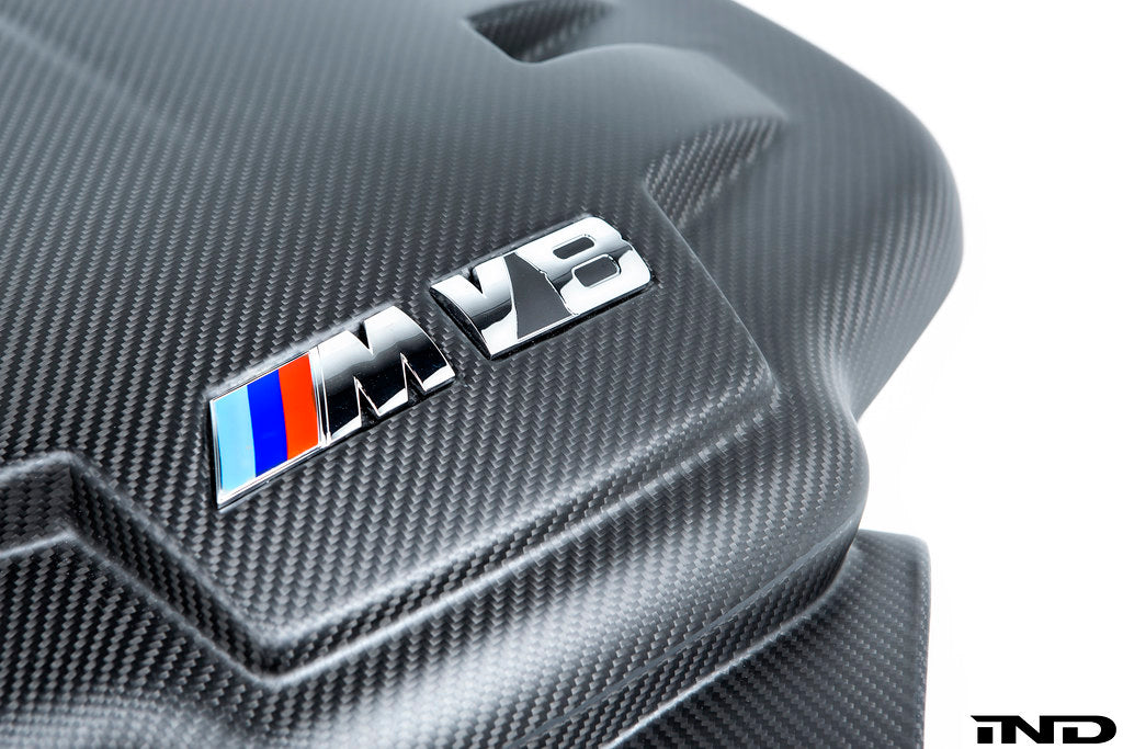 Close-up view of the BMW M V8 emblem installed on the Eventuri E9X M3 S65 Carbon Inlet Plenum in matte finish on a white background